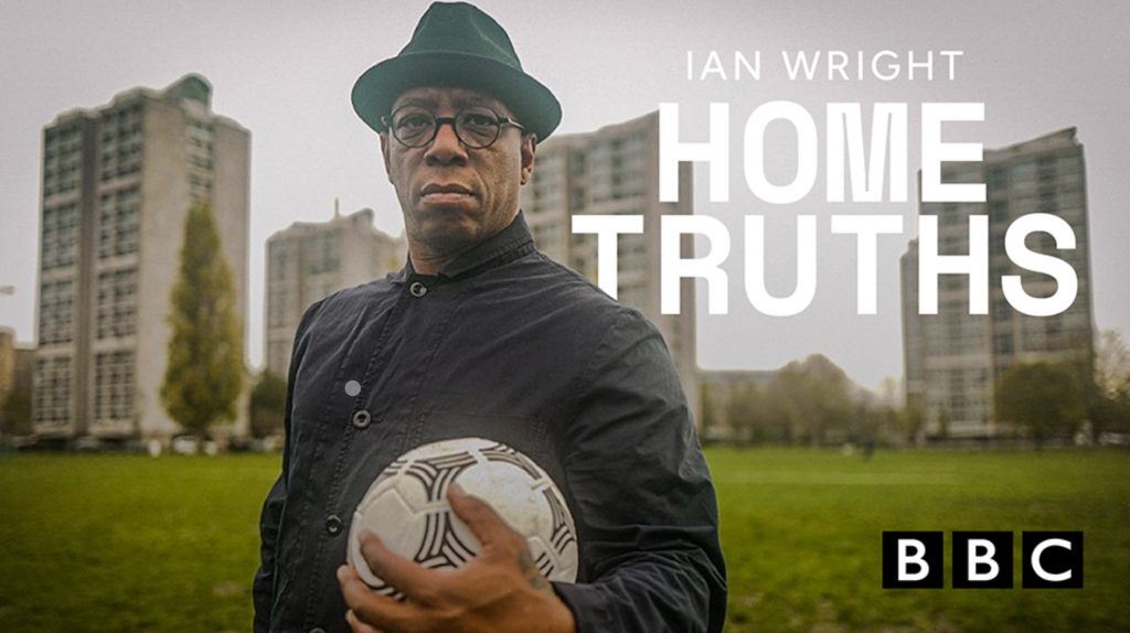 A man wearing a dark hat and jacket holds a football whilst standing in front of tall residential buildings. White text ‘Home Truths’ and the BBC logo appear next to him