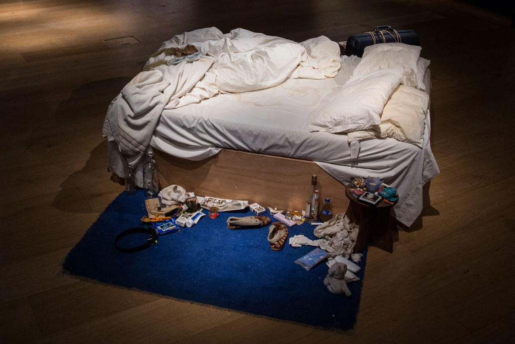 Emin’s installation showing an unmade bed surrounded by personal items like clothes, bottles, and slippers, representing the artist’s private space.