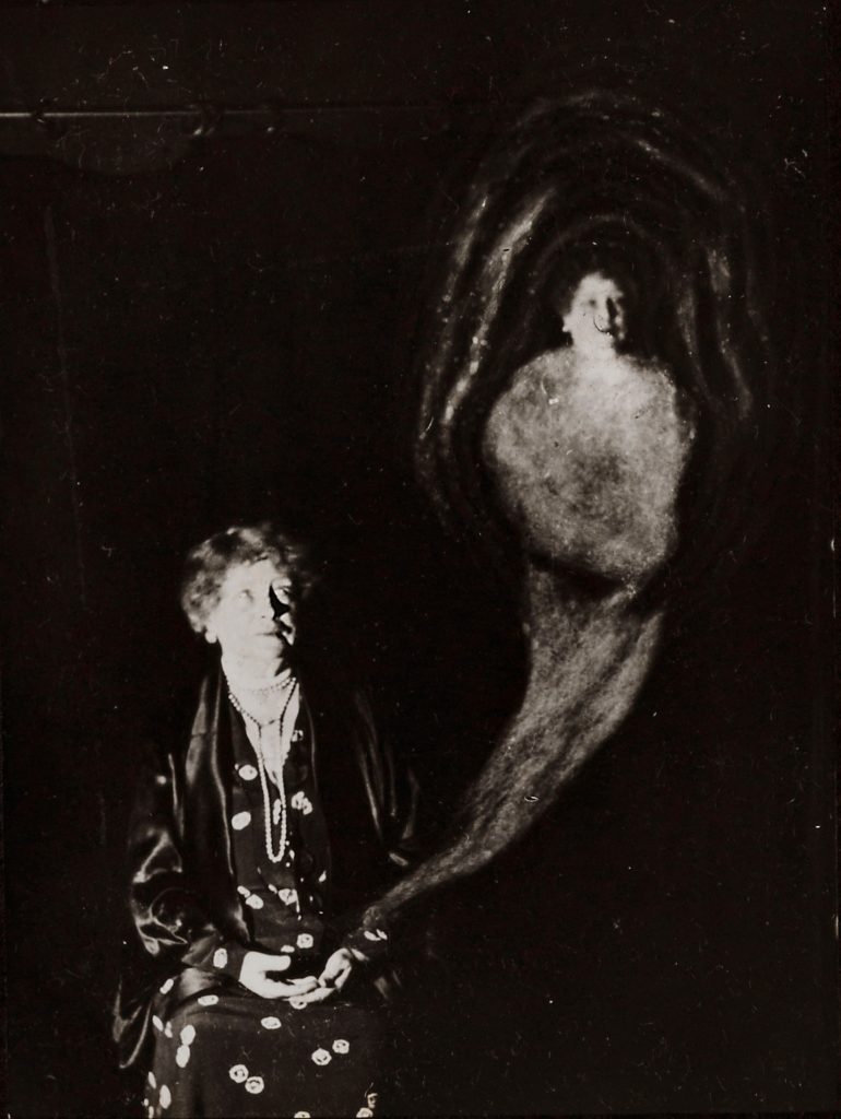 A seated woman in a dark room looks toward a faint, ghostly figure rising beside her, its hazy, human-like form appearing as a misty shape emerging from her hand