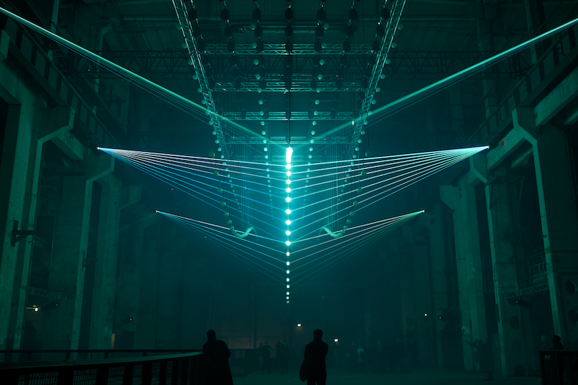 A blue coloured light installation of spherical shapes suspended by straight lines on either side projected near the ceiling of a dark warehouse-looking interior