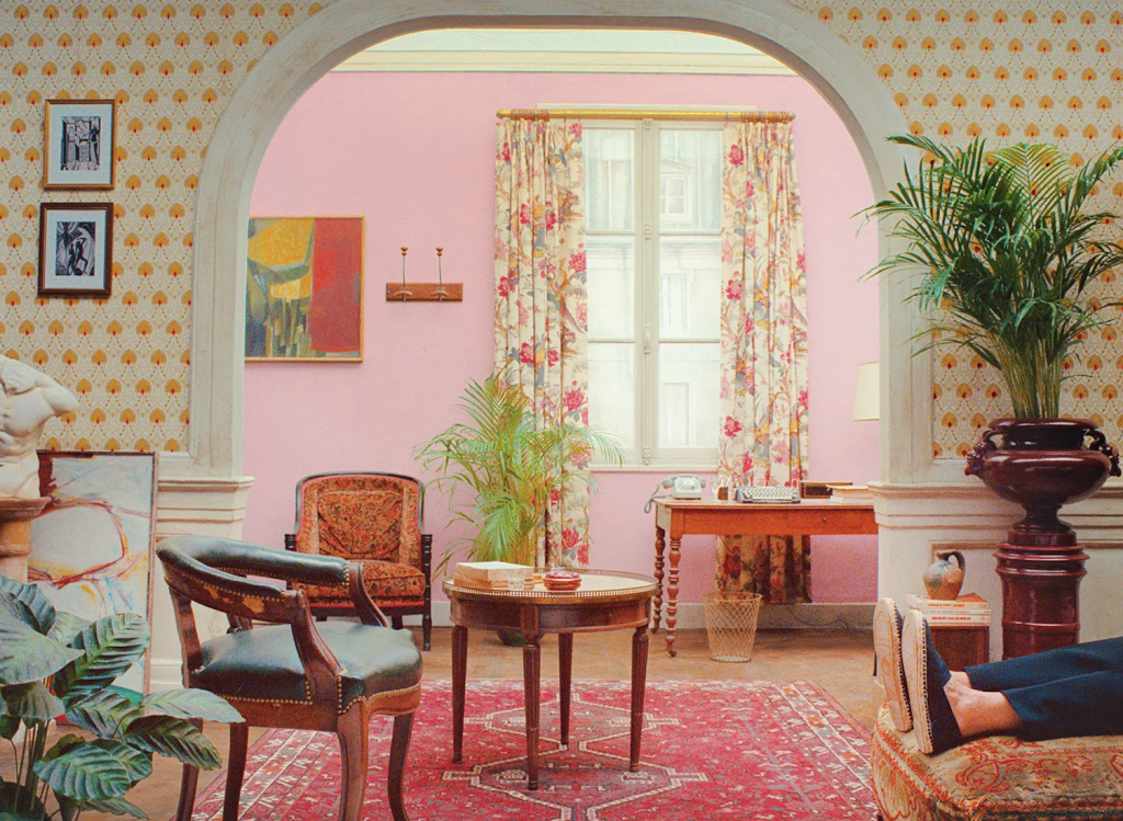 A pastel-toned interior framed by an arched doorway, featuring a pink wall with a central window dressed in floral curtains. Vintage wooden furniture, including chairs, a small round table, and a writing desk with a typewriter, is arranged neatly on a patterned red rug. Decorative elements such as framed artwork, houseplants, and patterned wallpaper create a warm, curated, mid-century aesthetic.
