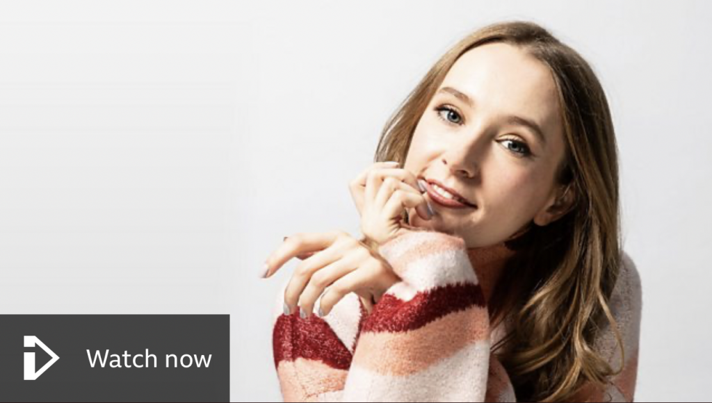 A young woman with long light brown hair leans forward with her chin resting on her hand, smiling softly at the camera. She wears a cozy striped sweater in red, pink, and cream tones. A ‘Watch now’ button with a play icon appears in the lower left corner against a light background.